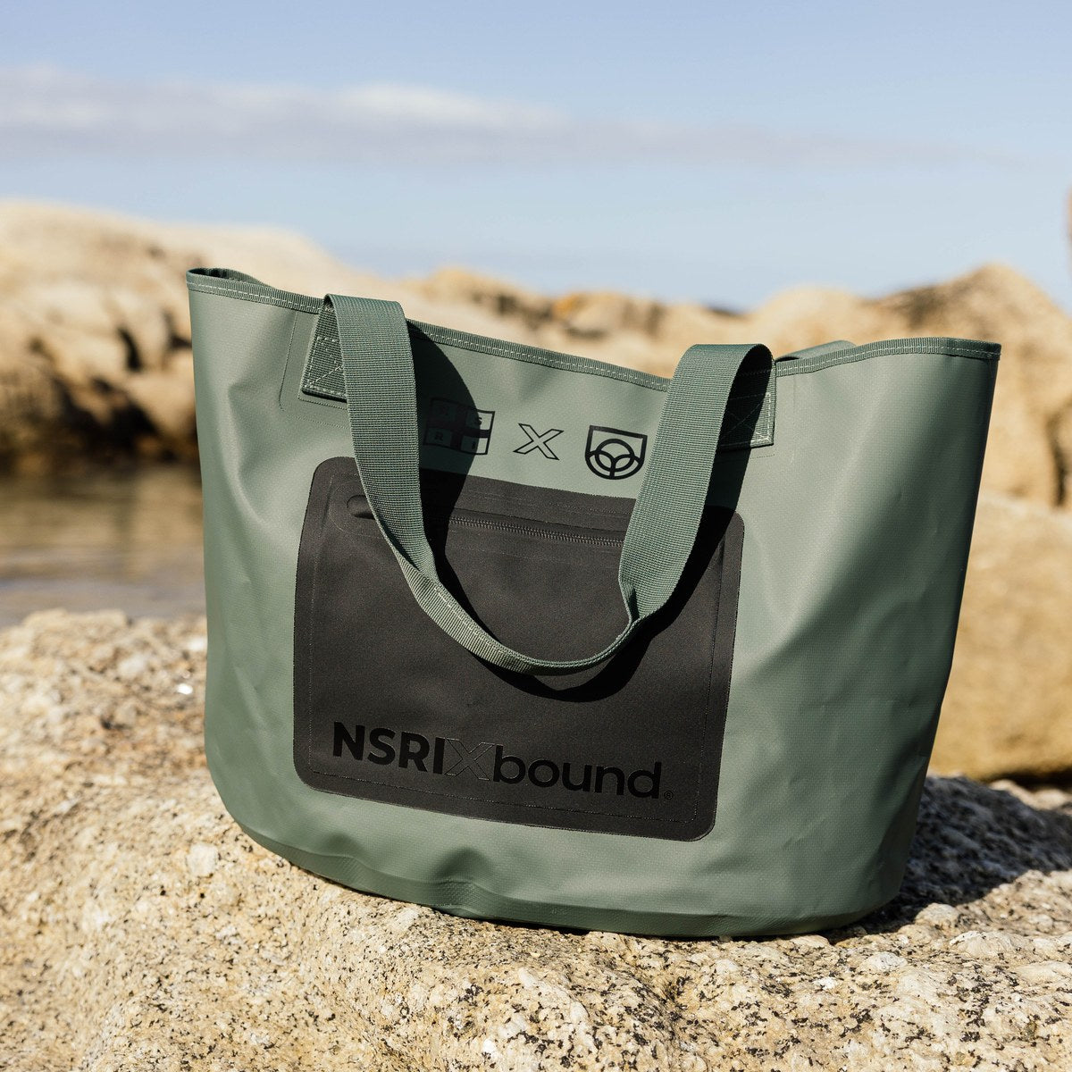 NSRI Tote in green standing on coastal rocks with the sea behind